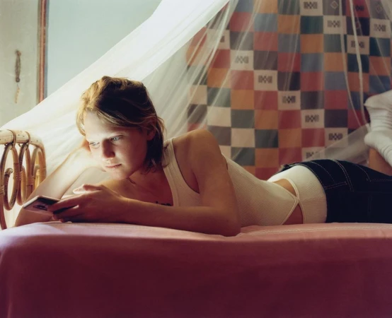Young woman with short blond hair reclines on a pink bed under a sheer canopy, holding a cell phone and looking at the screen.