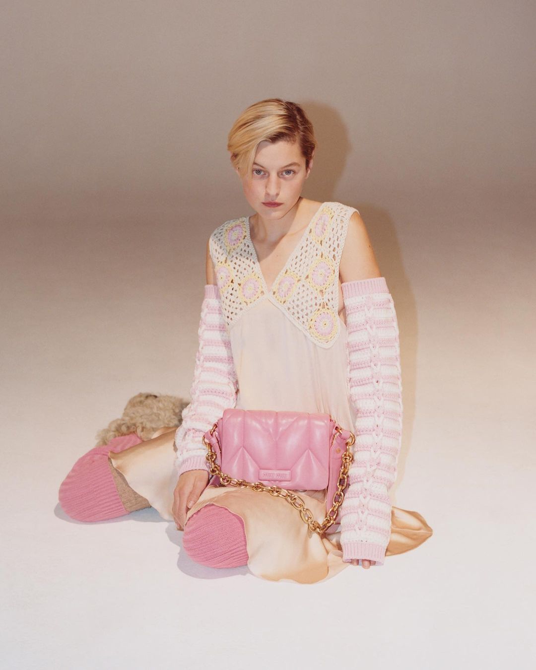 Fashion shot of a woman in a light-pink dress with crochet sleeves and a handbag, posed in a photo studio with a light-pink background.