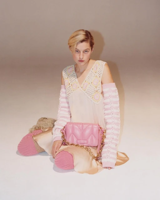 Fashion shot of a woman in a light-pink dress with crochet sleeves and a handbag, posed in a photo studio with a light-pink background.