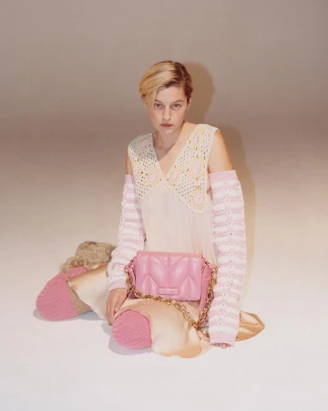 Fashion shot of a woman in a light-pink dress with crochet sleeves and a handbag, posed in a photo studio with a light-pink background.