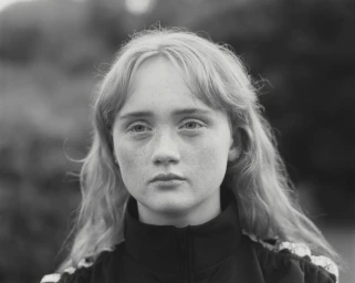 Portrait of a young woman with freckles and long blonde hair wearing a dark zip-up jacket, photographed by Julie Greve.