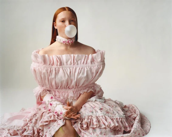 Red-haired woman in a pink off-the-shoulder dress blowing a bubblegum bubble against a white background