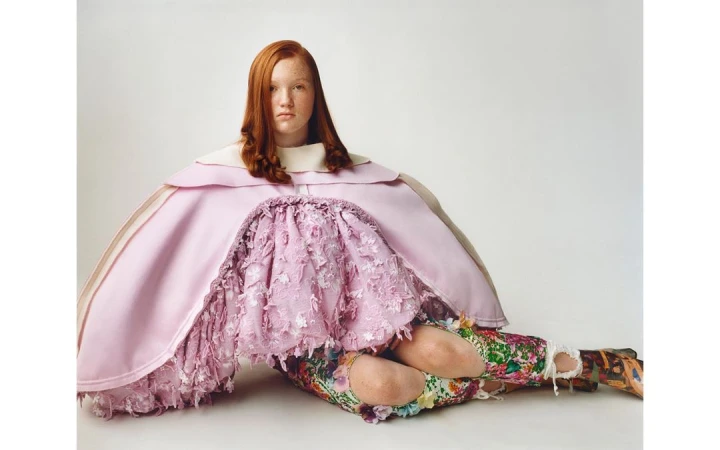 Red-haired model wearing a voluminous pink cape and floral skirt seated against a white background