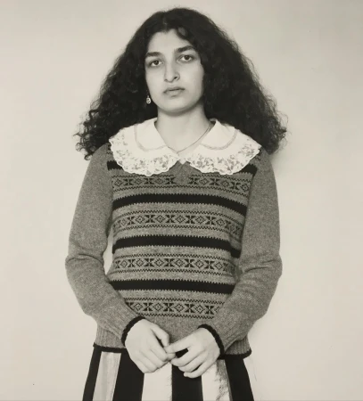 Portrait of a woman with dark curly hair wearing a patterned sweater and lace collar against a light background.