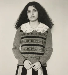 Portrait of a woman with dark curly hair wearing a patterned sweater and lace collar against a light background.