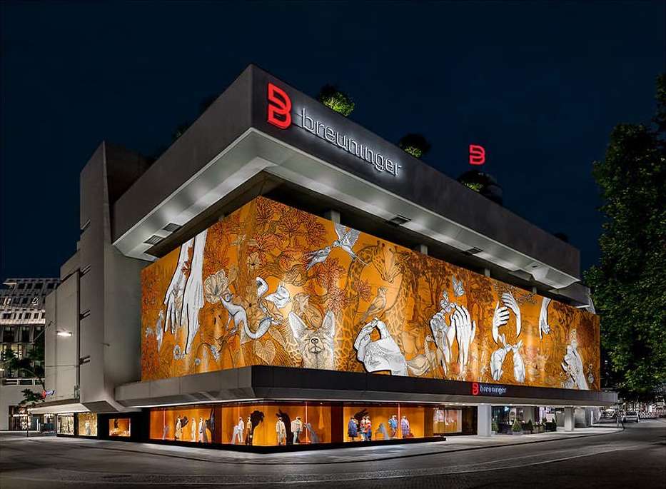 Illuminated multi-story Breuninger department store at night showcasing a large mural and window displays with mannequins and clothing.
