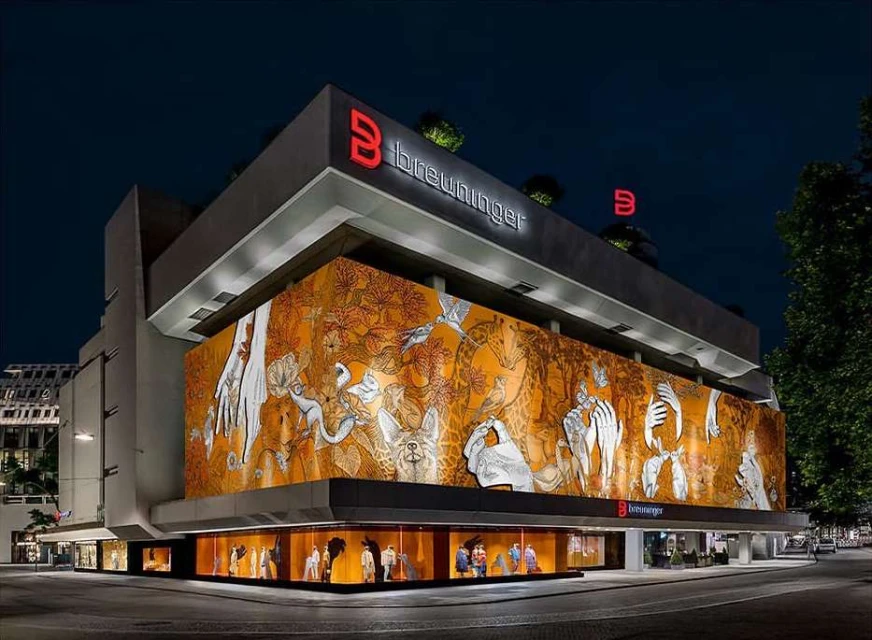 Illuminated multi-story Breuninger department store at night showcasing a large mural and window displays with mannequins and clothing.