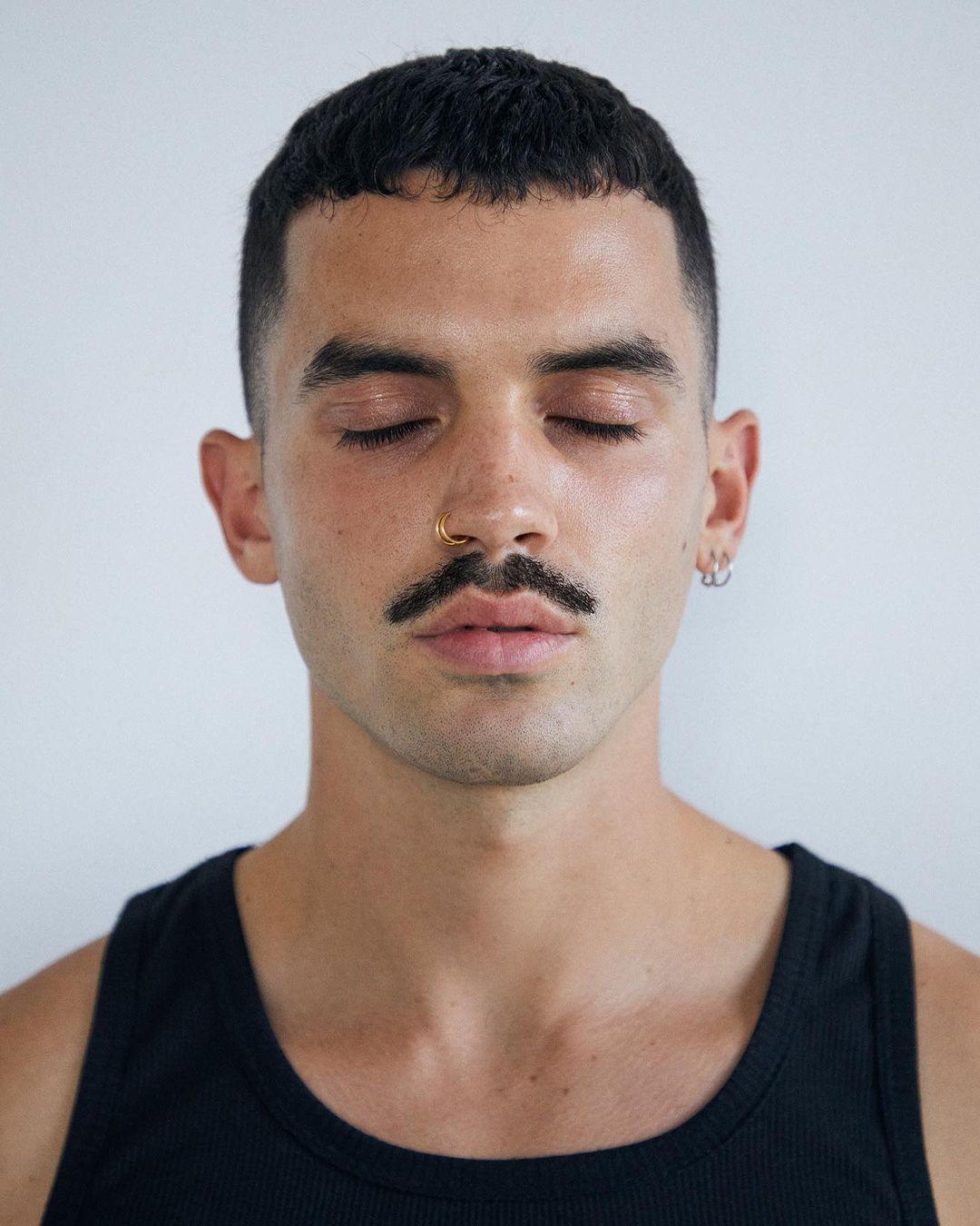 Portrait of a young adult man with a mustache and closed eyes, wearing a black tank top, against a neutral, light-colored backdrop.