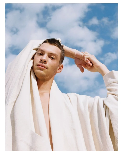 Young man poses outdoors in a white linen robe against a blue sky with clouds. He rests his hand behind his head in a relaxed pose.