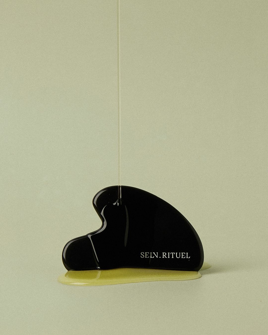 Black gua sha tool with liquid dripping on it against a light background. Focus on beauty treatment and skincare concept.
