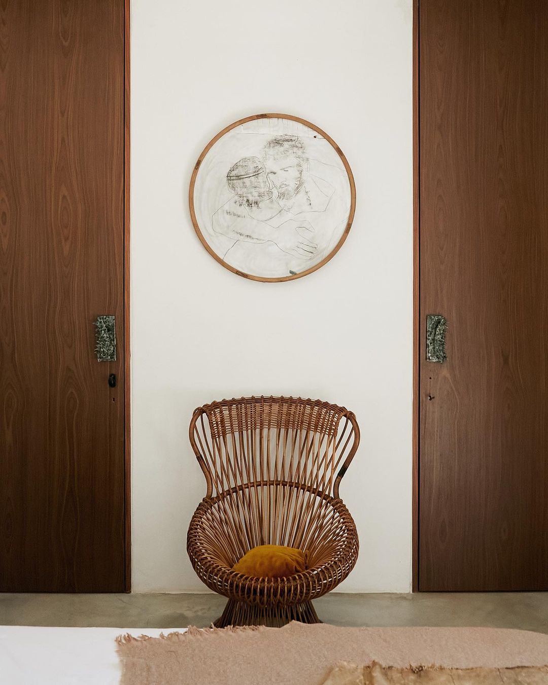 Interior view of a minimalist room with a wicker chair, a framed drawing, and paneled doors on either side of the white wall.