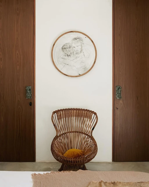 Interior view of a minimalist room with a wicker chair, a framed drawing, and paneled doors on either side of the white wall.