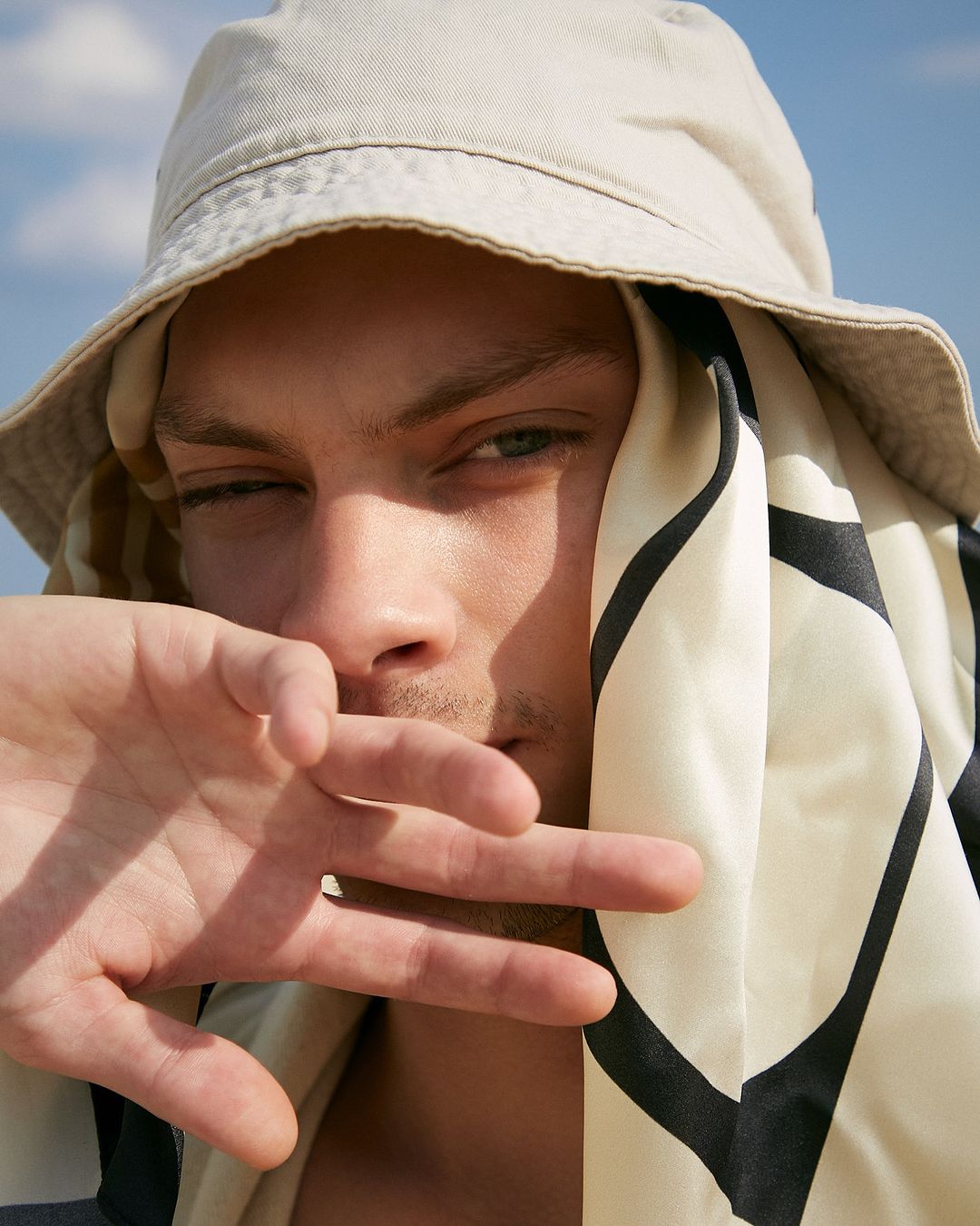 Close-up portrait of a man wearing a bucket hat and patterned headscarf while holding a hand near his face under clear blue sky.