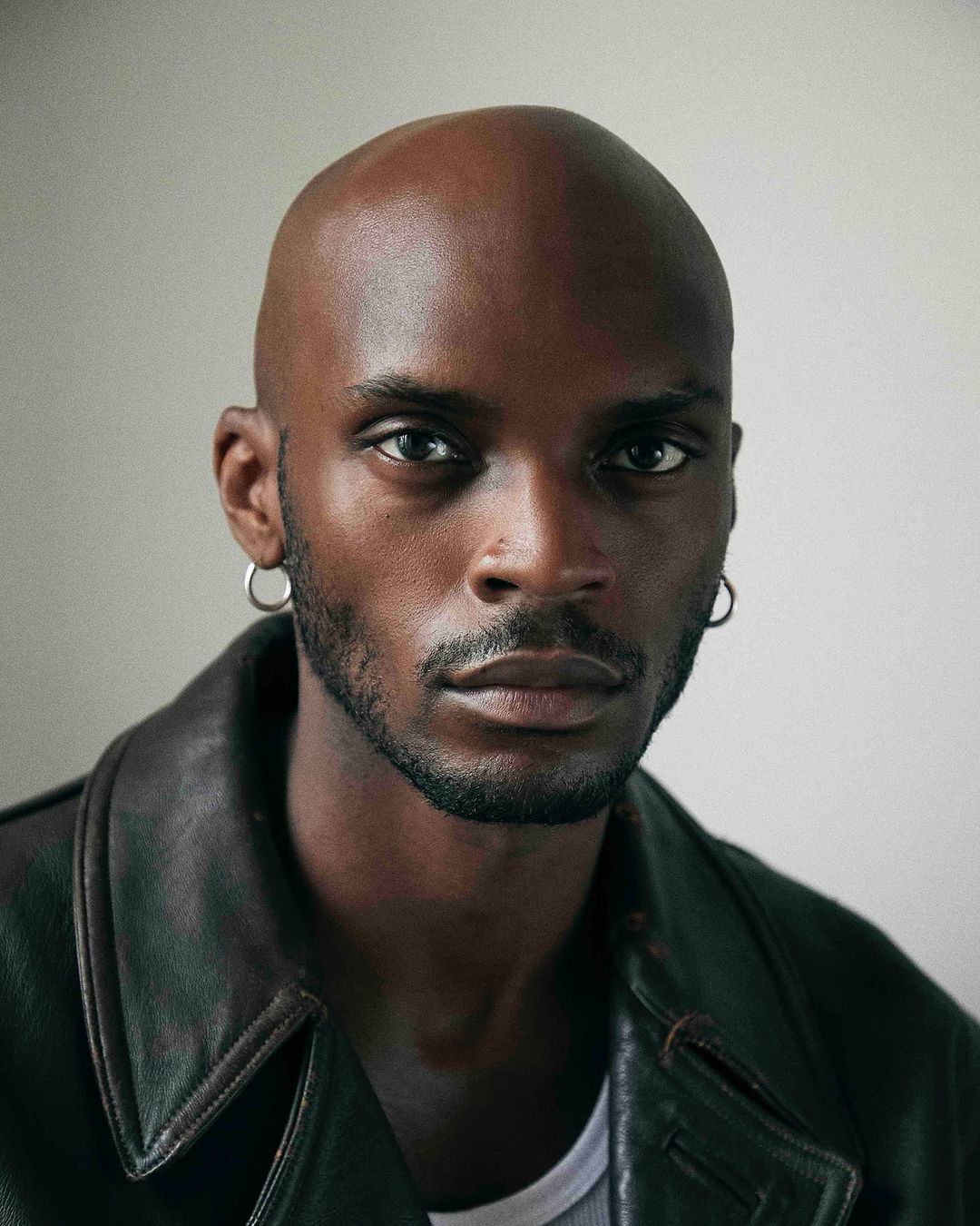 Portrait of a bald Black man with silver hoop earrings wearing a dark leather jacket over a white t-shirt.