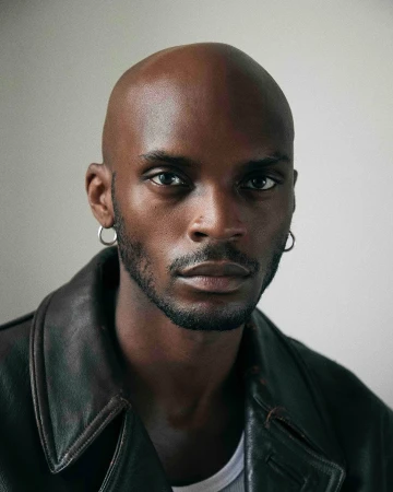 Portrait of a bald Black man with silver hoop earrings wearing a dark leather jacket over a white t-shirt.