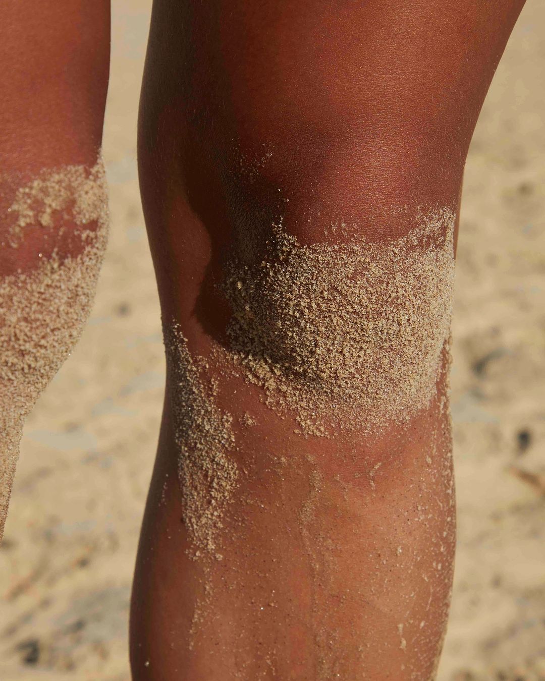 Close-up of tanned legs covered with sand on a beach, focusing on the texture of the skin and sand under bright, natural lighting.