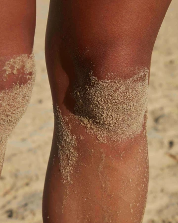 Close-up of tanned legs covered with sand on a beach, focusing on the texture of the skin and sand under bright, natural lighting.