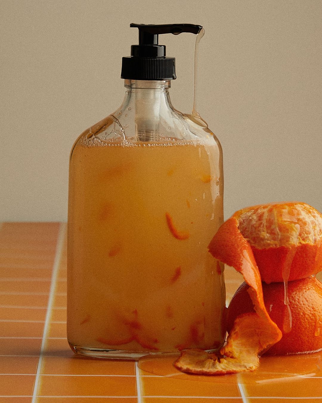 Still life photograph of a glass bottle filled with orange liquid and orange peel next to two whole oranges on a tiled surface.