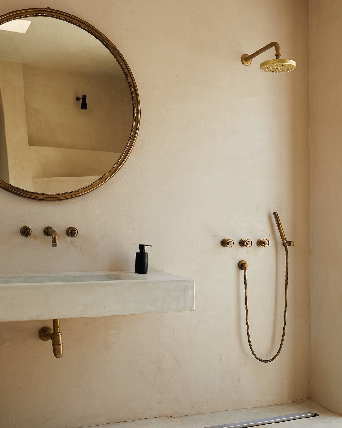 Minimalist bathroom interior with gold fixtures including a round mirror, shower head, and faucet set against a textured wall.