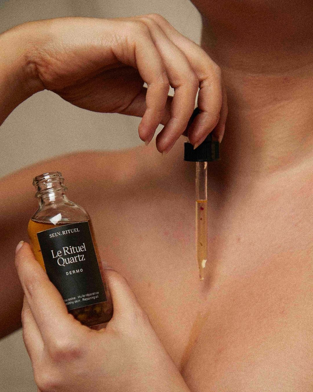 A woman applies serum to her chest with a glass dropper; a bottle of skincare product is held in her other hand; natural light.