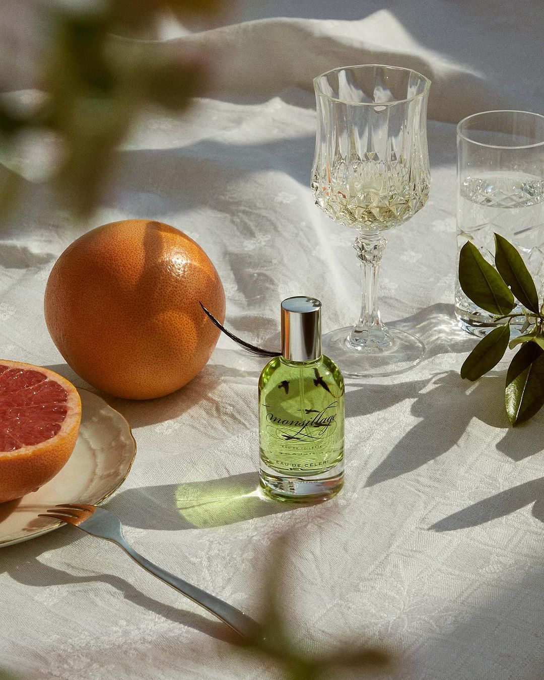 Close-up of a bottle of green perfume next to grapefruit slices, a glass of white wine, and a fork on a floral tablecloth.