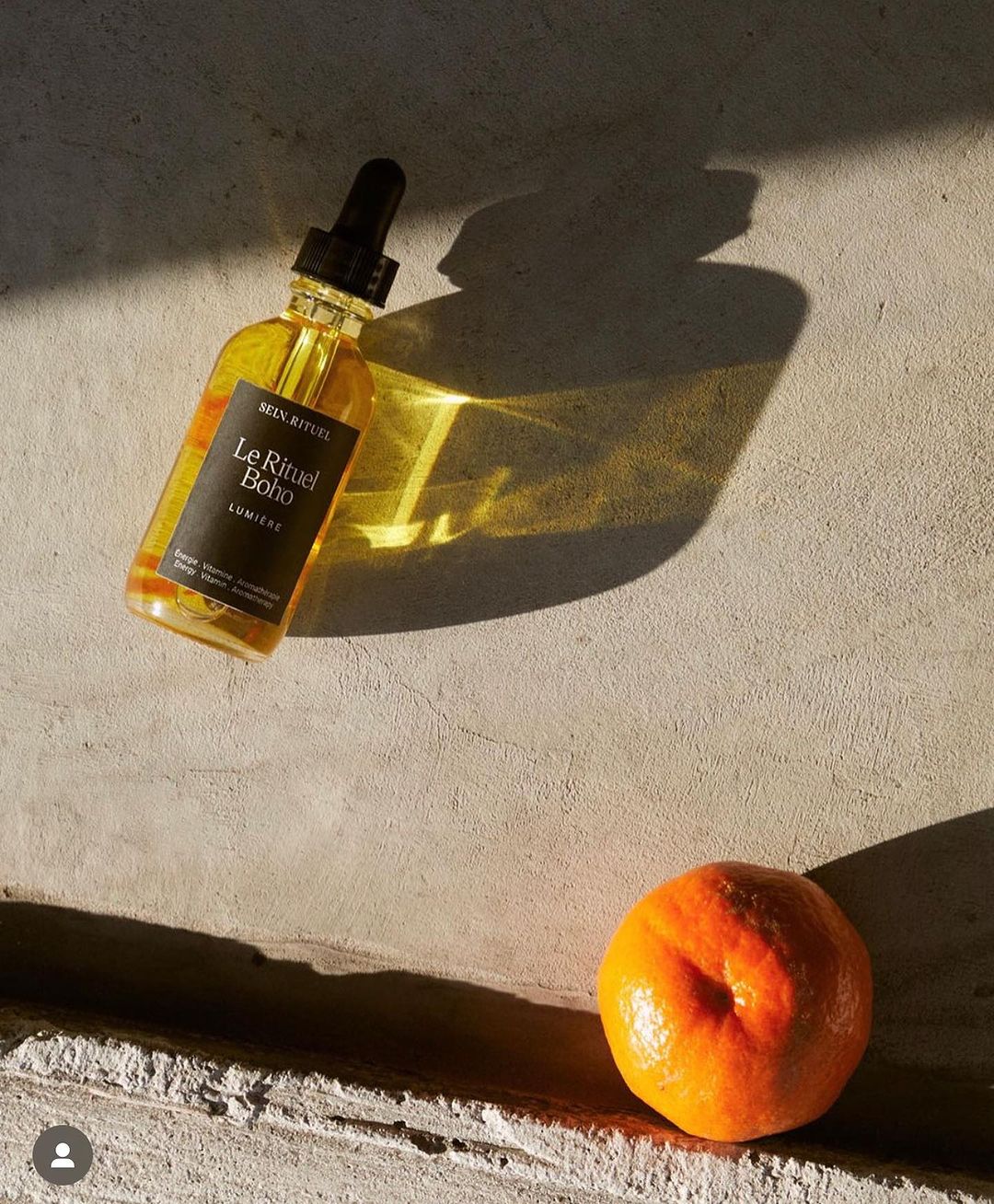 A glass bottle of cosmetic oil with dropper cap rests on a concrete surface with an orange citrus fruit casting a sunlit shadow.