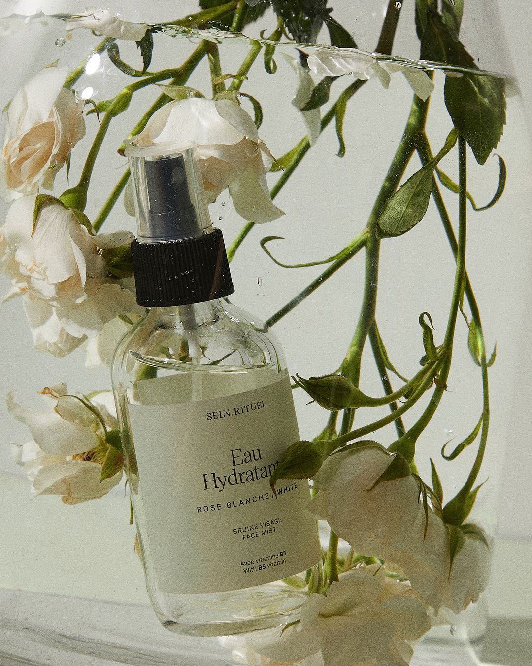 Beauty product shot: clear spray bottle with a black nozzle is surrounded by white roses submerged in water against a white background.