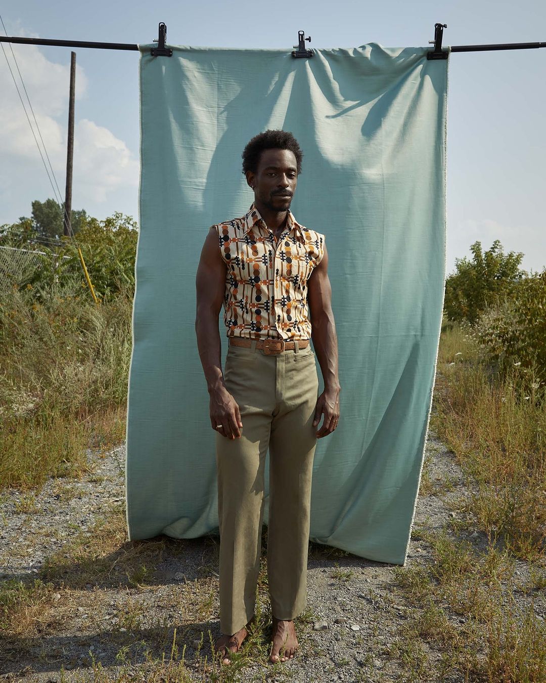 A black male model poses outdoors in sleeveless printed shirt and khaki pants in front of a draped blue fabric backdrop on a sunny day.