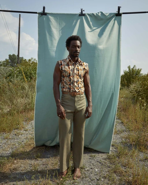 A black male model poses outdoors in sleeveless printed shirt and khaki pants in front of a draped blue fabric backdrop on a sunny day.