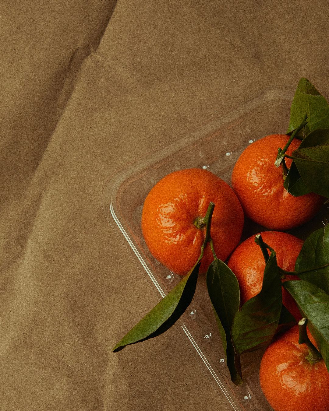 Overhead shot of fresh tangerines on a clear plastic tray, set against a textured brown paper background. Bright, natural lighting.
