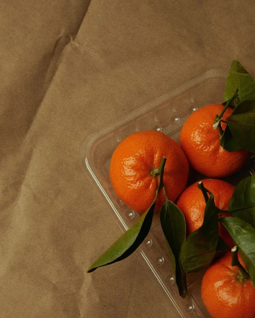Overhead shot of fresh tangerines on a clear plastic tray, set against a textured brown paper background. Bright, natural lighting.