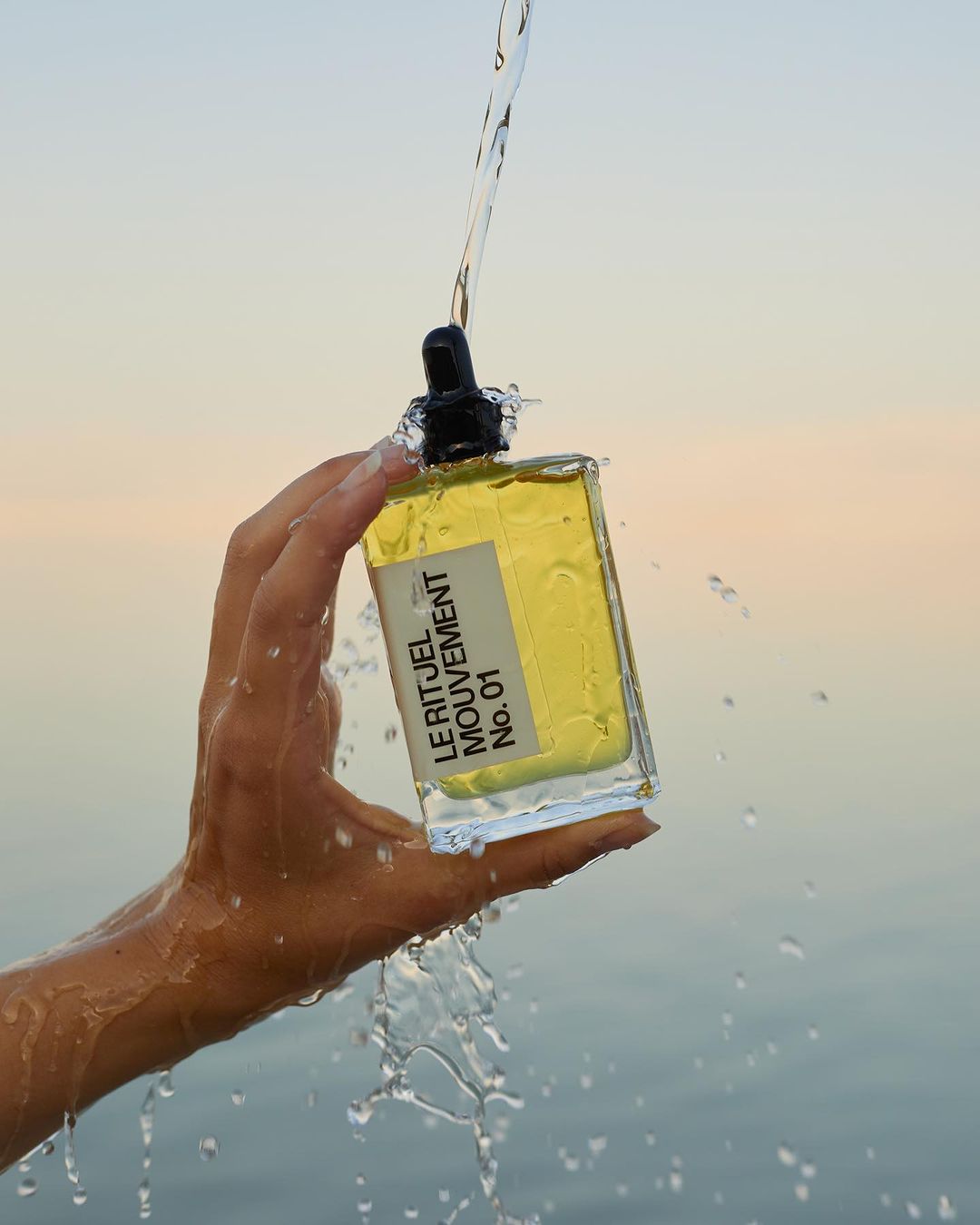 Woman's hand holding a bottle of Le Rituel Mouvement No. 01, with water running down the bottle, against a soft sky background.