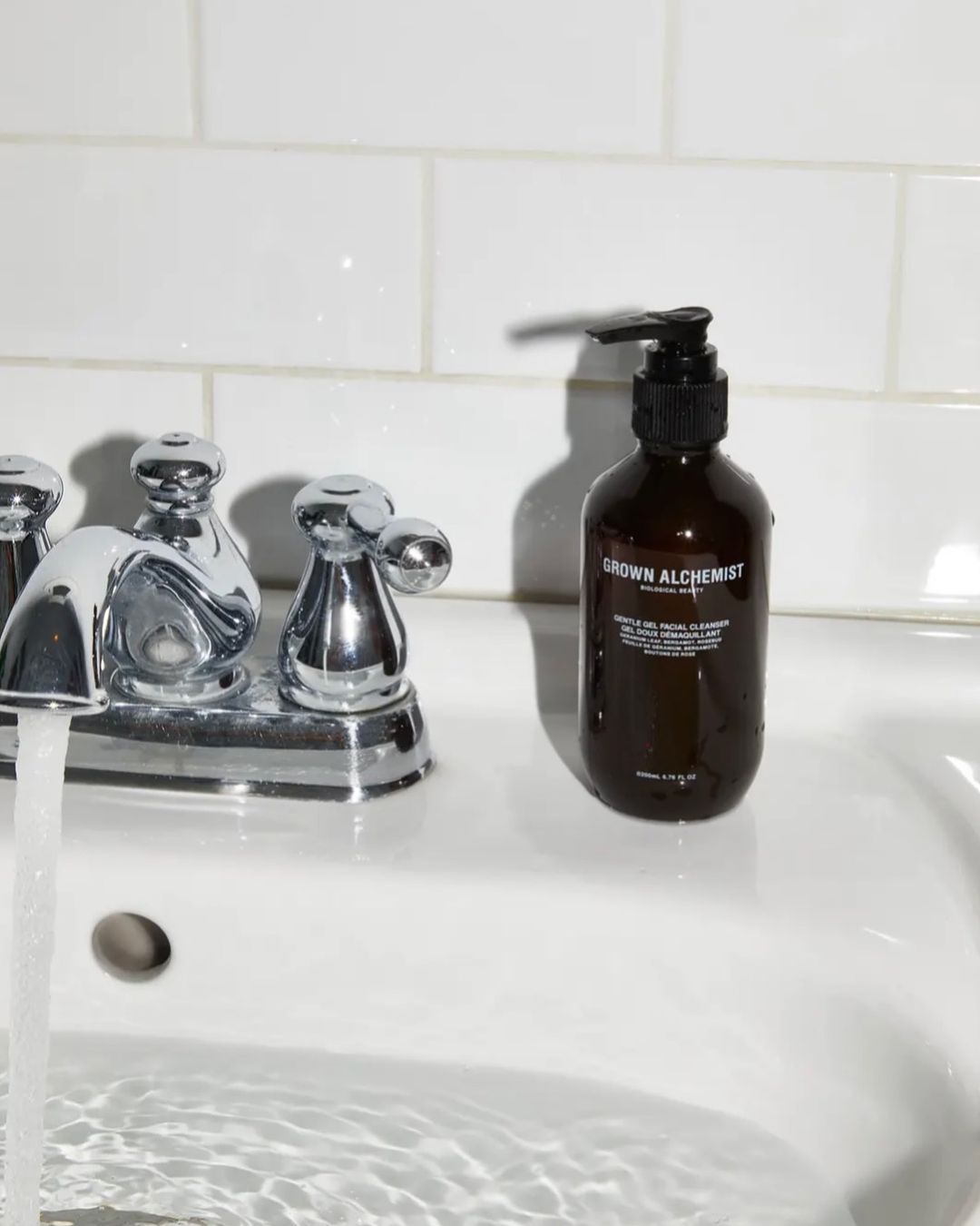 Brown glass pump bottle on a white porcelain sink with chrome faucet; water is running in a bathroom with white tiled walls.
