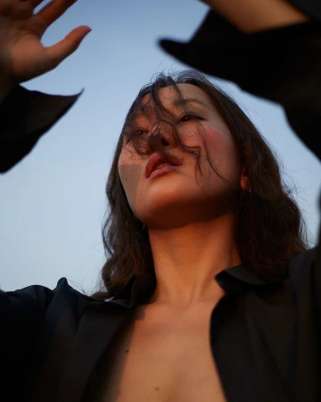 Low angle portrait of a woman wearing a black collared shirt with hair blowing across her face against a clear blue sky