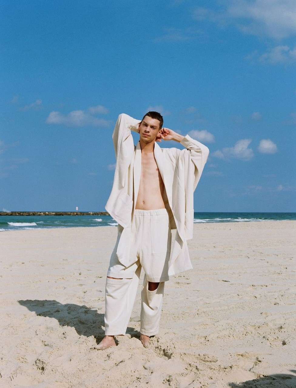 Man in a white jacket and pants stands on a beach with blue sky, ocean, and light clouds, posing with hands behind his head.