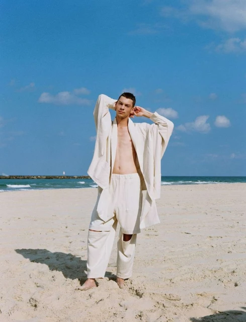 Man in a white jacket and pants stands on a beach with blue sky, ocean, and light clouds, posing with hands behind his head.