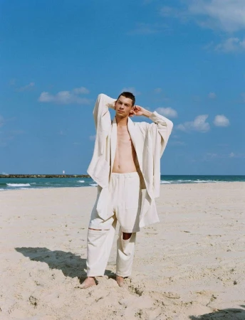 Man in a white jacket and pants stands on a beach with blue sky, ocean, and light clouds, posing with hands behind his head.