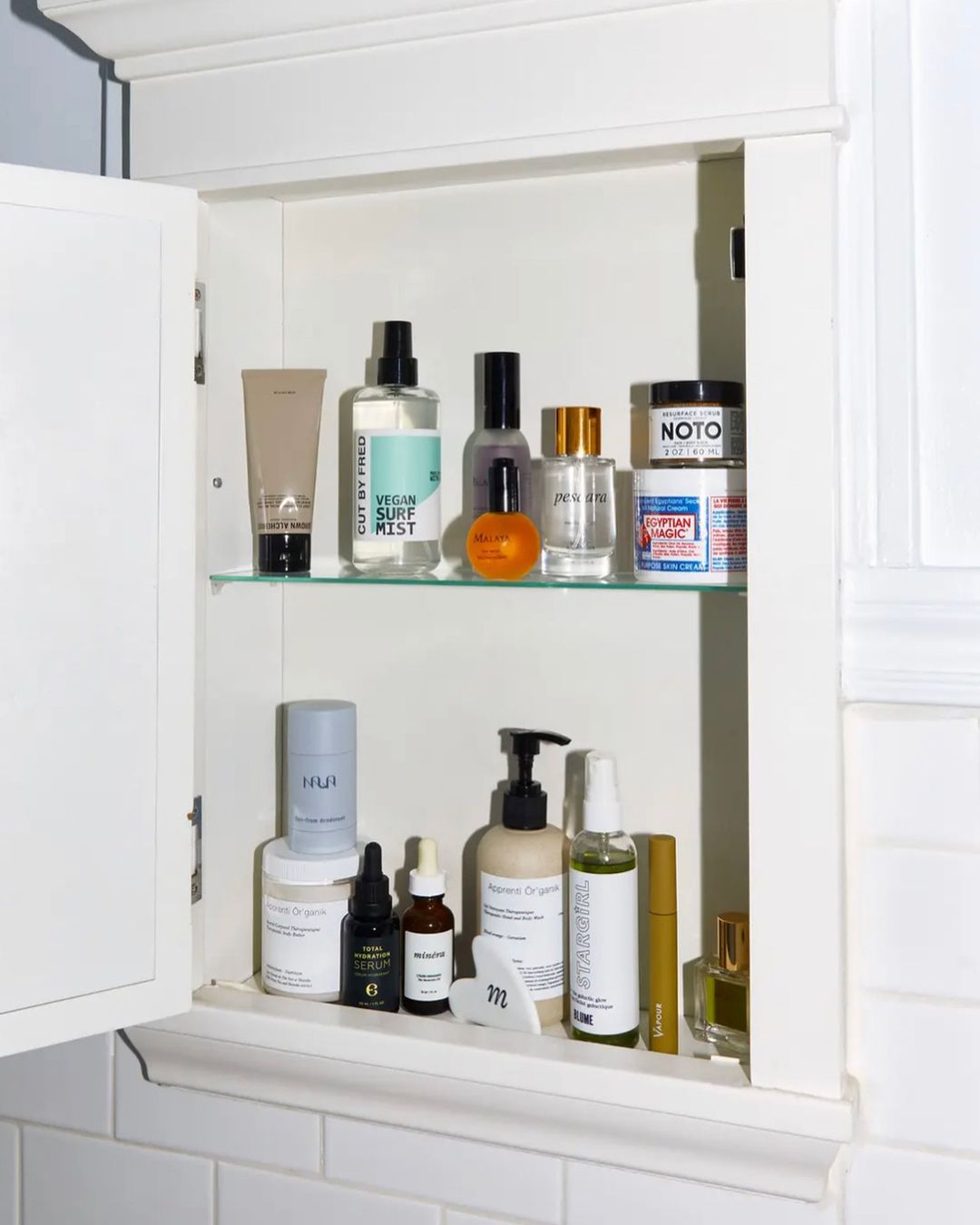 Bathroom cabinet with various beauty and skincare products including creams, serums, sprays, and lotions arranged on glass shelves.