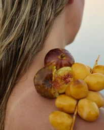 Close-up portrait of a woman with wet blonde hair holding fresh figs and yellow dates on her shoulder.