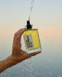 Hand holding a yellow glass perfume bottle as water streams over it against a sunset sky.