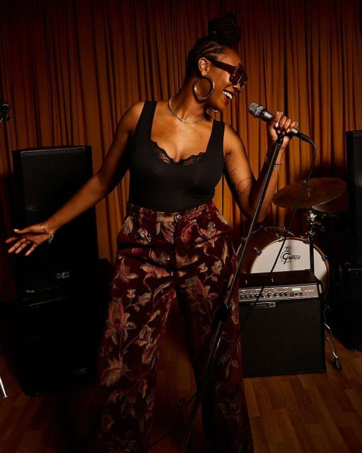 A Black female musician sings into a microphone on stage with a drum kit and speakers. She is wearing sunglasses and patterned pants.