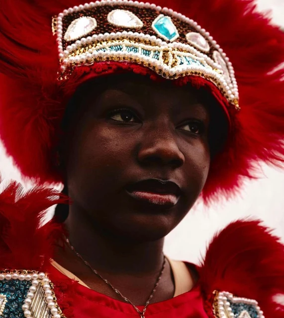 Close-up of an adult female wearing an elaborate red feathered headdress with beads, looking off to the side, indoors with diffused light.