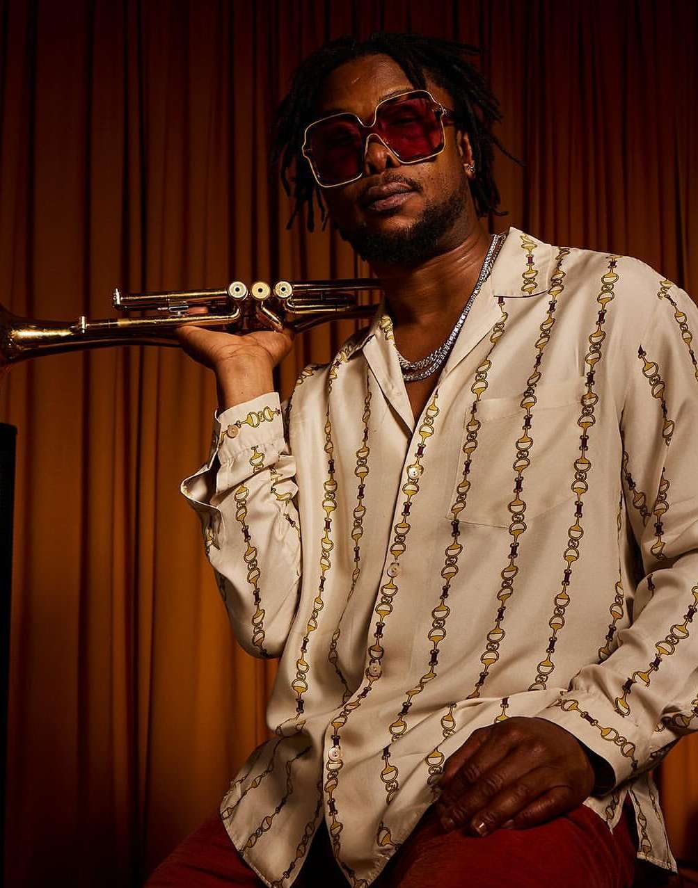 Portrait of a Black male musician in patterned shirt holding a trumpet against a dark backdrop, wearing tinted sunglasses and jewelry.