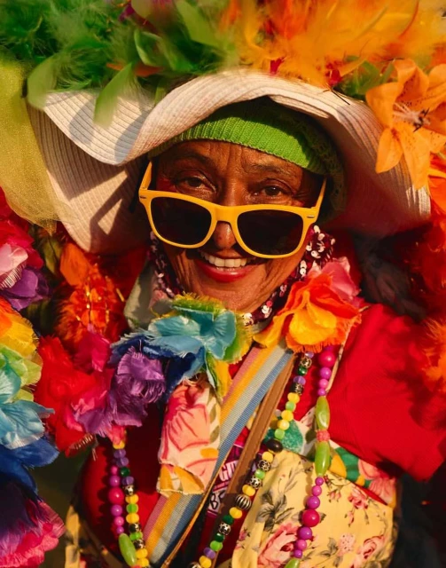 Close-up of a woman in bright yellow sunglasses, a wide-brimmed hat with feathers, and colorful beaded necklaces smiling at the camera.