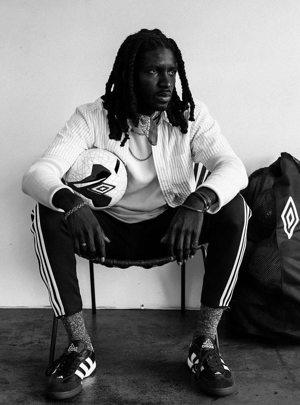 A black and white portrait of a Black male athlete with dreadlocks sitting with a soccer ball, wearing athletic pants, jacket, and shoes.