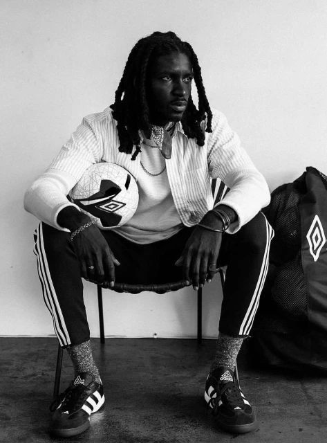 A black and white portrait of a Black male athlete with dreadlocks sitting with a soccer ball, wearing athletic pants, jacket, and shoes.