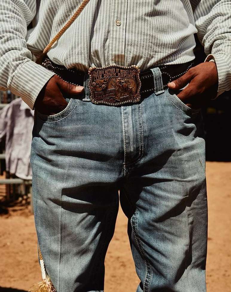 A medium studio shot focuses on an adult person wearing a cowboy belt buckle, striped button shirt, and blue jeans, with hands in pockets.