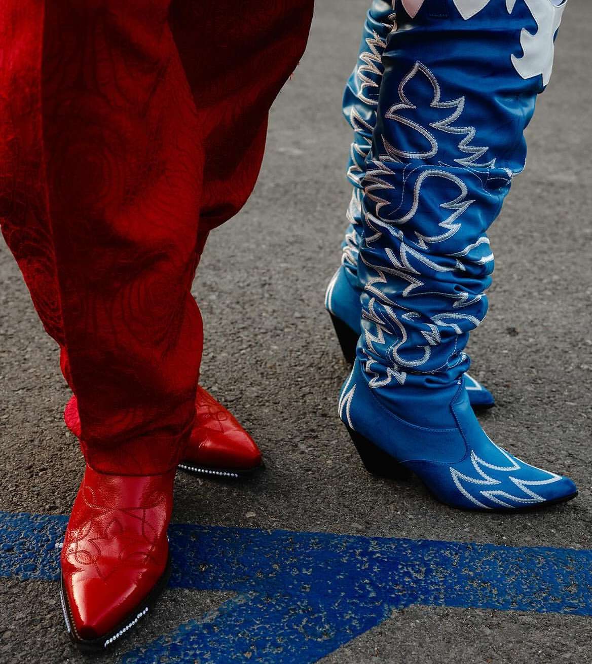 Close-up of two people wearing brightly colored cowboy boots, one red and one blue with white designs, standing on a gray asphalt surface.