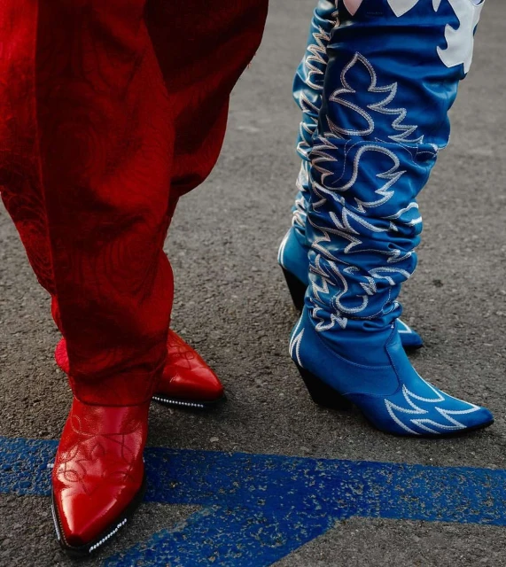 Close-up of two people wearing brightly colored cowboy boots, one red and one blue with white designs, standing on a gray asphalt surface.