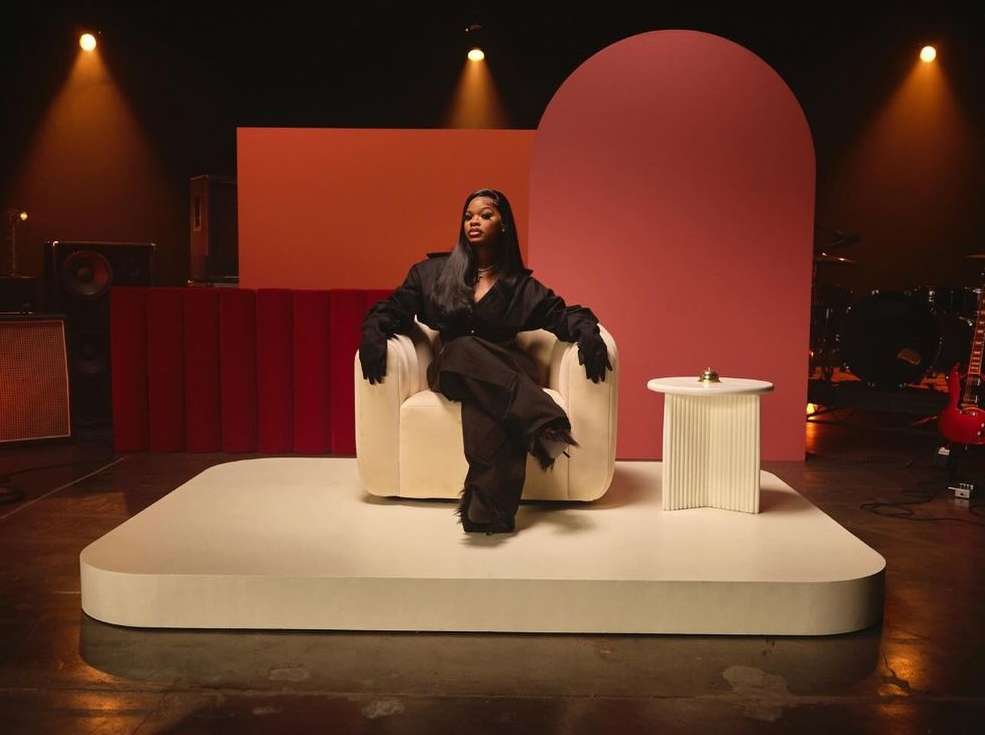 A woman with long dark hair sits in a cream-colored armchair on a raised platform, wearing a black suit, gloves, and black pants.